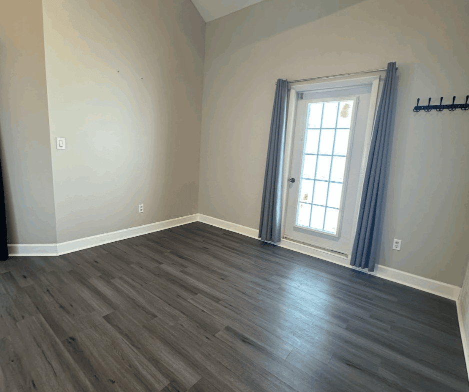 Bright empty room with large window, neutral walls, and gray wood flooring.